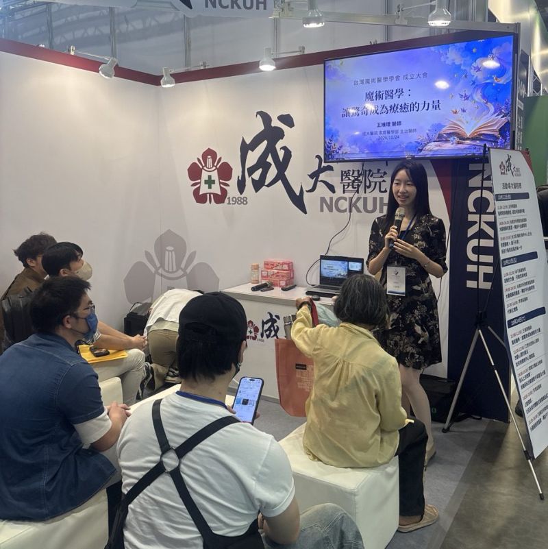 王維理醫師站在成大醫院攤位前手持麥克風演講，投影幕顯示講題「魔術醫學：讓驚奇成為療癒的力量」，民眾坐在前方聆聽